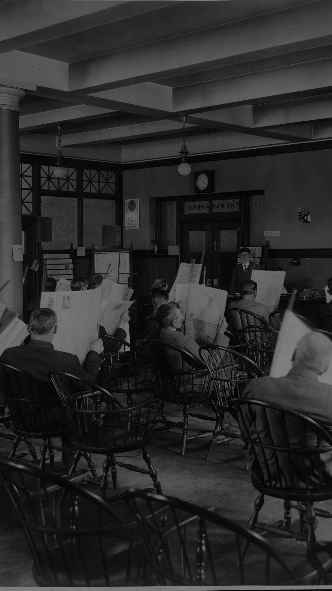 Rows of men in wooden chairs reading newspapers in a library reading room, Seattle, around 1910.